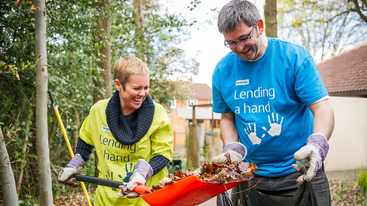 A smiling man and woman from Skanska putting leaves into a bag as part of a volunteering day